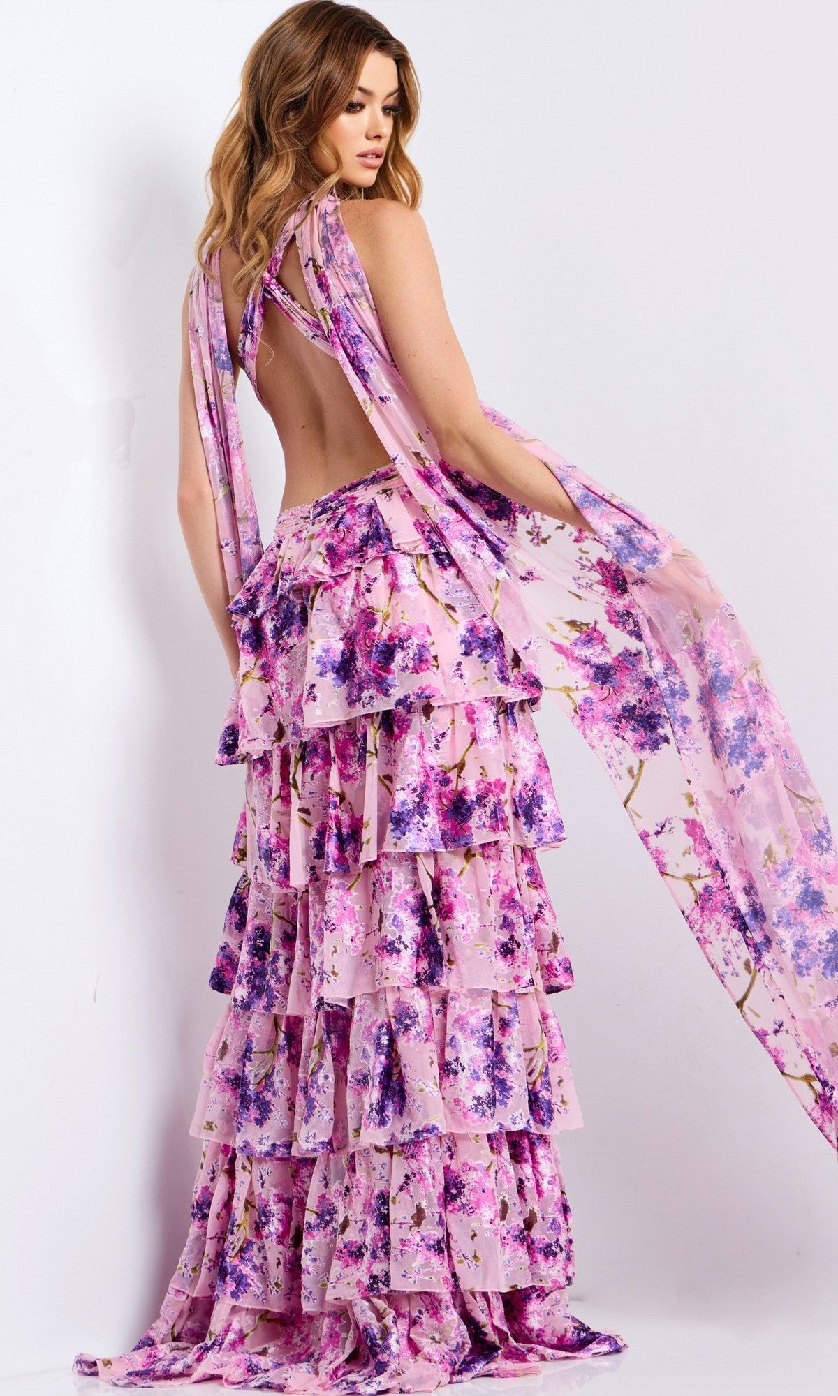 A woman models the Jovani 48234 Long Prom Dress, a sleeveless, backless, floor-length gown with pink and purple floral patterns and layered ruffles, against a plain white background.